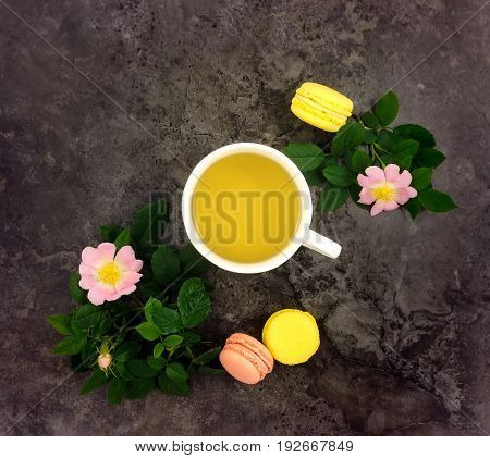 Summer bright composition of cup of herbal tea three macarons dogrose branches and flowers on dark marble table. Flat lay top view