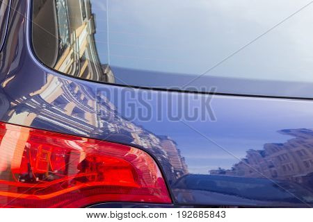 Fragment of the back of the modern blue car with with tail lamps and brake lights and reflection of the neighboring houses on it