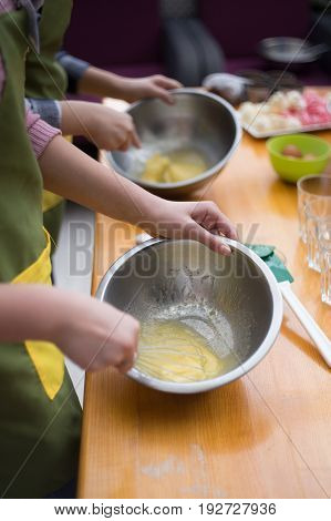 Cookery scene with female hands beating eggs with eggbeater in metal bowls on light brown table at the background of cooking ingredients. Shallow dof. Focus on hand.