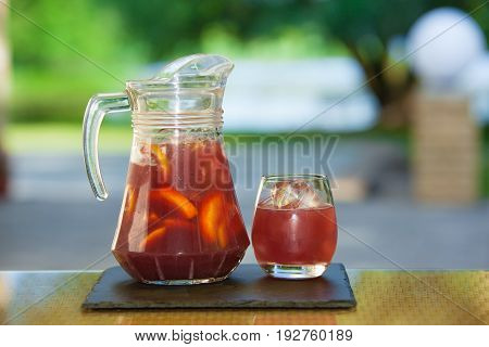 Jug of non-alcoholic drink and a glass of ice on a table with a blurred background.
