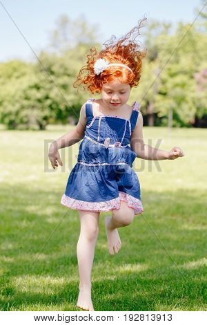 Portrait of cute adorable little red-haired Caucasian girl child in blue dress jumping on grass in meadow park outside funny candid moment happy lifestyle childhood concept