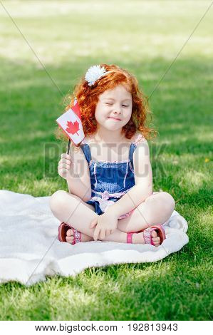 Portrait of cute little red-haired Caucasian girl child holding Canadian flag with red maple leaf sitting on grass in park outside celebrating Canada Day anniversary