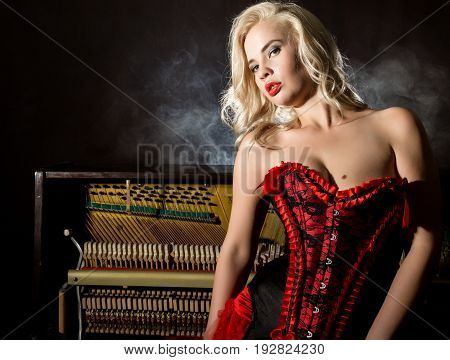 Young beautiful girl in red corset standing near the piano. Vintage style beautiful woman. Old fashioned makeup and retro finger wave hairstyle.