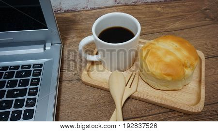 Cup of black coffee  with bun on wooden plate. Spoon & fork and laptop computer.
Breakfast or coffee break time.