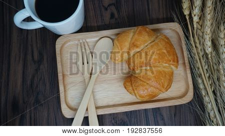 A Cup of coffee , croissant  on wooden plate with forkand wheat on table