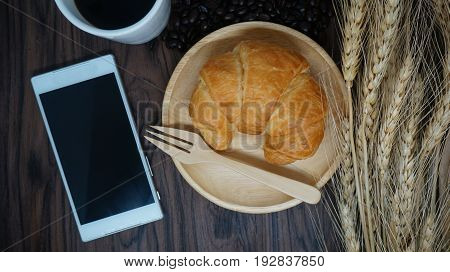 Croissant  on wooden plate with fork, coffee cup,mobile phone and wheat on table