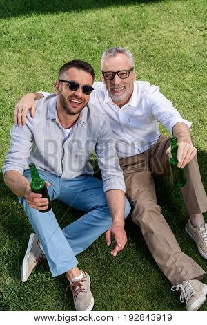 Happy Father And Adult Son Drinkig Beer And Sitting On Green Grass