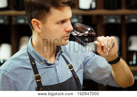Sommelier smelling flavor of red wine in bokal on background of shelves with bottles in cellar. Male appreciating color, quality and sediments of drink. Professional degustation expert in winemaking.