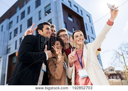 business, education and corporate concept - international group of people with conference badges taking selfie by smartphone on city street
