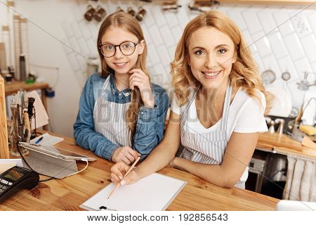 Inspired owners. Lovely mother and daughter sitting at the coffee bar counter and creating a new business plan for their recently opened cafe, both of them smiling at the camera