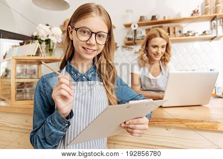 Up for new tasks. Beautiful teenage girl smiling at the camera and being about to put down a new task in her to-do list on the sheet holder while her mother working on the laptop in the background