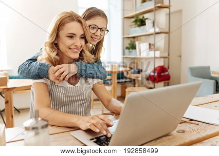 Loving hugs. Pleasant cheerful daughter hugging her mother from behind and peeking at the laptop screen while her mother typing a letter