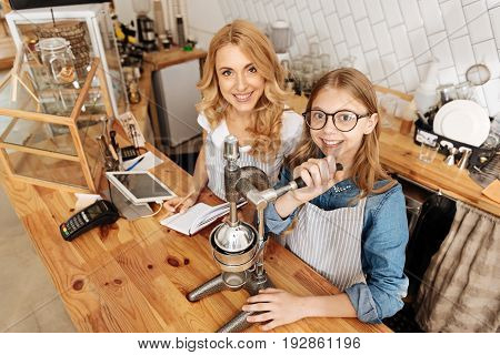 Successful attempts. Pretty teenage girl looking happy while successfully making fine coffee under careful supervision of her mother, both of them smiling at the camera