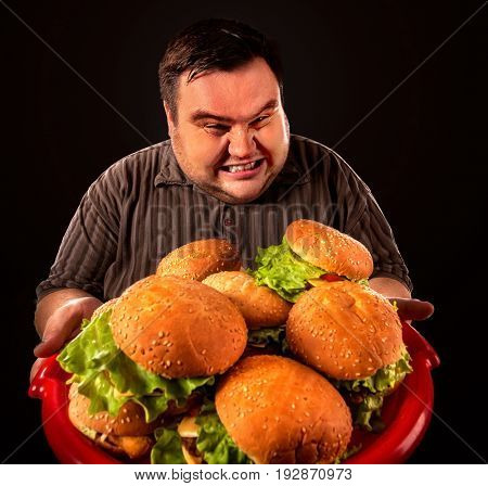 Fat man eating fast food hamberger and carries treat for friends on tray. Overweight person drops tray of food. Junk meal leads to obesity. Person regularly overeats concept on black background.