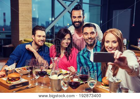 leisure,technology, friendship, people and holidays concept - happy friends having dinner and taking selfie by smartphone at restaurant