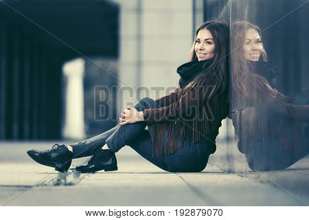 Happy young woman sitting at the wall in city street. Stylish fashion model in leather fringe suede jacket and blue jeans