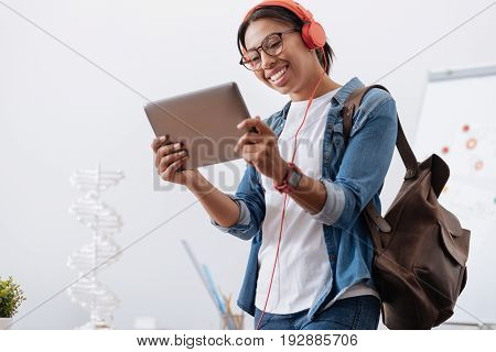 Modern igeneration. Happy delighted young student wearing headphones and using a tablet while holding a bag