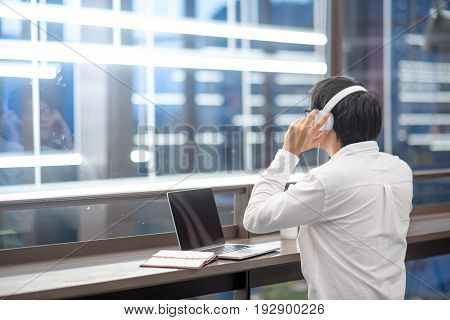 Young Asian man dressed in casual style listening to music while working with his laptop. Digital nomad in co working space modern IT lifestyle with work life balance concept.