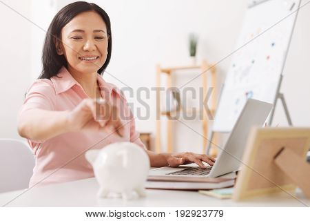 Save for future. Cheerful asian woamn sitting at the table and working in the office while putting coins into a piggy bank