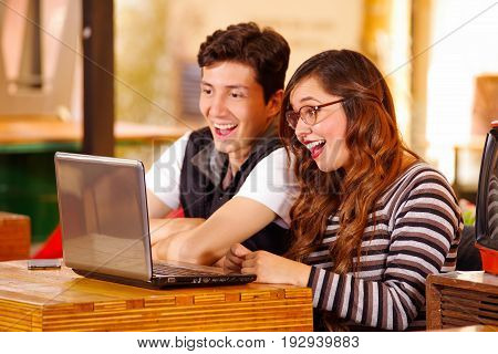 Happy beautiful couple at the bar spending time together, while she is working in her computer on a wooden table wearing glasses, while he is using his cellphone.