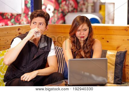 Smiling happy couple at the bar spending time together, man drinking a bottle of water while his girlfriend is working in her computer.