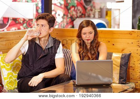 Smiling happy couple at the bar spending time together, man drinking a bottle of water while his girlfriend is working in her computer.