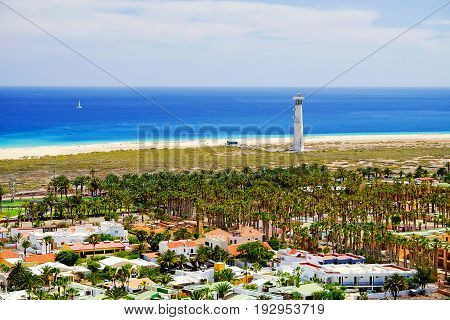 Aerial view on Morro Jable with homes for holidays with green palm trees around a lighthouse the beach with golden sand Playa de Morro Jable or Marorral and white sailboat in the Atlantic Ocean. Canary Island Fuerteventura Spain.