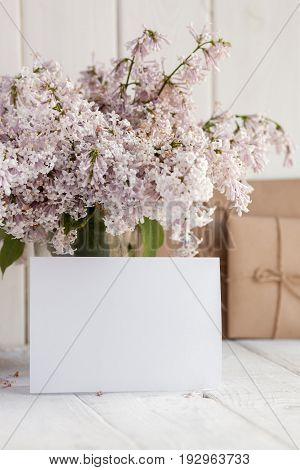 Blank white greeting card with lilac flowers bouquet and envelope on white wooden background. mock up.