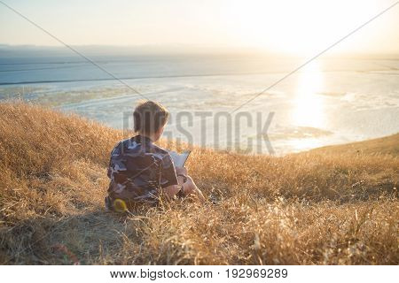 Boy reading book in yellow grass high in the hills, watching beautiful sunset above Bay Area, rear view