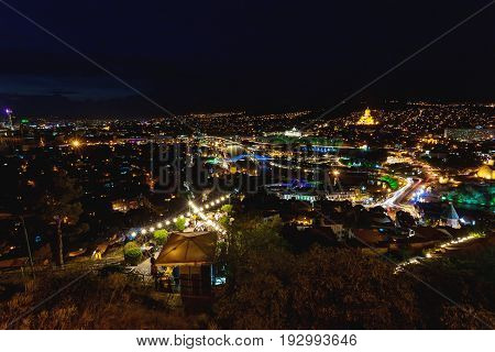 Sunset panorama view of Tbilisi capital of Georgia country from Narikala fortress. Famous landmarks with illumination. Tourists have dinner in restaurant with old fashioned light bulbs.