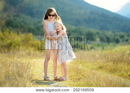 Two Cute Young Sisters Laughing And Hugging On Warm And Sunny Summer Day During Family Vacations In