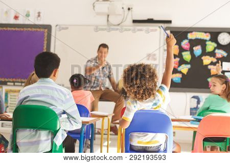 Rear view of school kids raising hand while teacher asking question in classroom at school