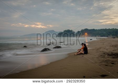 Happy Couple Enjoying Their Moments On Karang Hawu Beach, West Java, Indonesia With An Orange Sunset