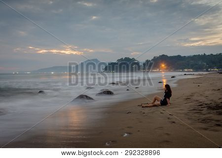 A Woman Is Sitting And Enjoying The Moment On Karang Hawu Beach, West Java, Indonesia With An Orange
