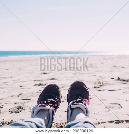 Feet Of Young Woman In Sport Footwear And Jeans Relaxing After Hiking On Sand Beach. Square Pov. Adv