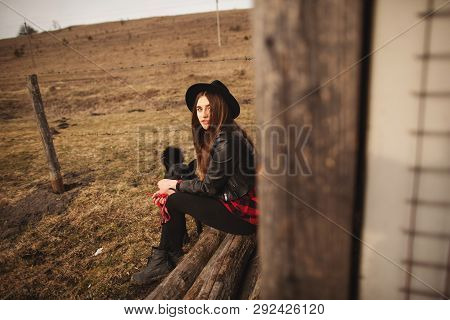 Happy Young Woman Sitting With Her Black Dog In Fron Of Old Wooden House
