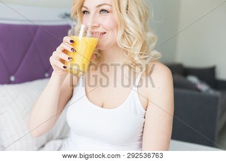 Attractive Blonde Woman Sits On The Edge Of The Bed At Home And Holds A Stocker With Orange Juice In