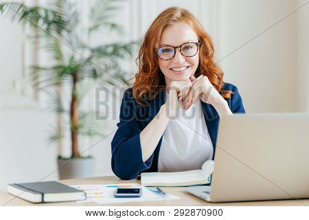 Pleased Cheerful Red Haird Female Economist Develops Financial Startup Project, Poses In Office Inte