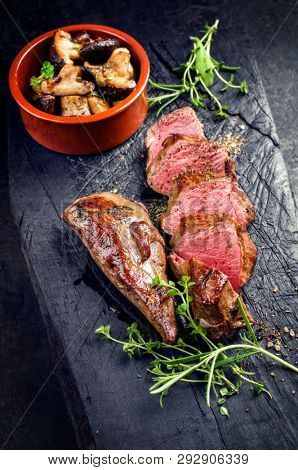 Traditional barbecue aged sliced venison sirloin with mushroom and herbs as closeup on a carbonized old board 