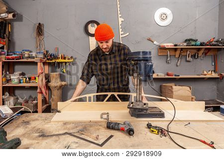 A Man Carpenter In A Hat And A Shirt  Is Carving A Wooden Board On A Large Drilling Machine In A Wor