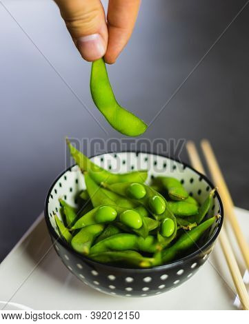 Hand Picking Up An Edamame Bean From A Bowl. Fresh Green Soybeans In A Bowl With Chopsticks.