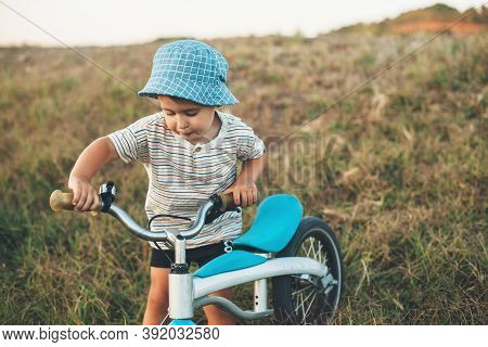 Caucasian Small Boy Walking With A Bike On A Field Wearing A Blue Hat In A Summer Day
