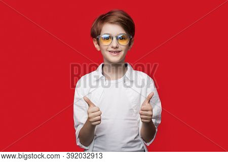 Young Caucasian Boy With Glasses Is Gesturing The Like Sign On A Red Studio Wall Smiling At Camera