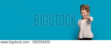 Smiling Cute Girl Is Gesturing The Stop Sign On A Blue Studio Wall With Free Space Wearing A Dress