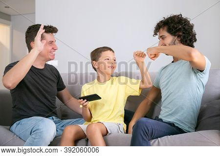Happy Boy With Cellphone Making Fist Bumping Gesture With Two Cheerful Dads. Fathers And Son Playing