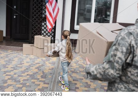 Back View Of Girl Carrying Cardboard Box Near House With Blurred Father On Foreground, Stock Image