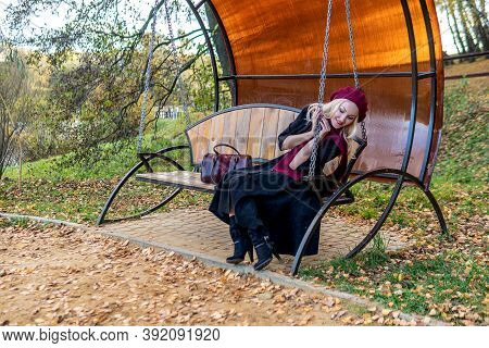 A Woman Sits On A Garden Swing, Forged In A Burgundy Coat And Biret, An Adult Looks At The Camera, I