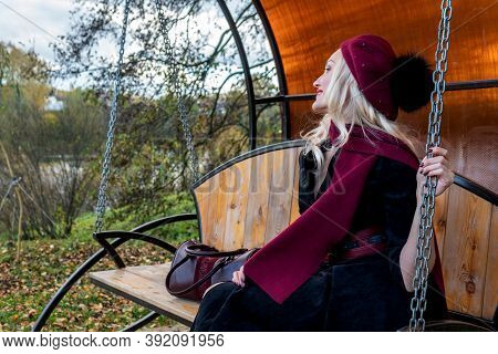 A Cheerful Girl Sits On A Garden Swing, Forged In A Burgundy Coat And Biret, An Adult, In The Fall A