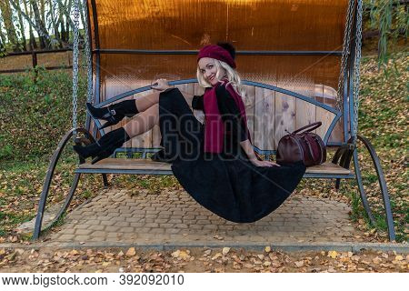 A Beautiful Woman Sits On A Garden Swing, Forged In A Burgundy Coat And Biret, An Adult Smiles At Th
