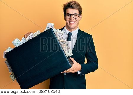 Handsome caucasian business man holding briefcase full of dollars smiling with a happy and cool smile on face. showing teeth. 